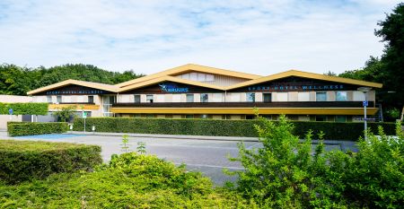 Das Sporthotel Bruurs ist ein modernes zweist&ouml;ckiges Geb&auml;ude mit gelben D&auml;chern und Wellness-Schildern, umgeben von gr&uuml;nen Hecken und B&auml;umen in Baarle-Nassau unter einem teilweise bew&ouml;lkten Himmel - ideal f&uuml;r ein Trainingslager.