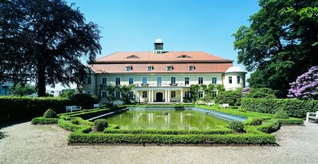 Das Hotel Schloss Schweinsburg ist ein prächtiges historisches Herrenhaus mit rotem Ziegeldach und großen Fenstern, das einen Blick auf gepflegte Gärten mit einem rechteckigen Teich, Hecken, Bänken, hohen Bäumen und nahegelegene Sportplätze bietet, auf de