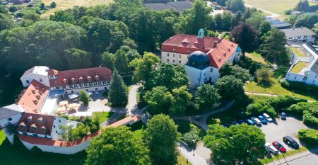 Das Hotel Schloss Schweinsburg ist ein prächtiges historisches Herrenhaus mit rotem Ziegeldach und großen Fenstern, das einen Blick auf gepflegte Gärten mit einem rechteckigen Teich, Hecken, Bänken, hohen Bäumen und nahegelegene Sportplätze bietet, auf de