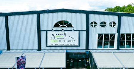 Das Hotel Stadt Mühlhausen*** besticht durch seine weiße Fassade im Industrial-Stil mit großen Fenstern. Ein Schild über dem Eingang weist auf das Hotel hin, das von Bäumen und einem bewölkten Himmel umgeben ist – ideal für Gäste des Trainingslagers oder