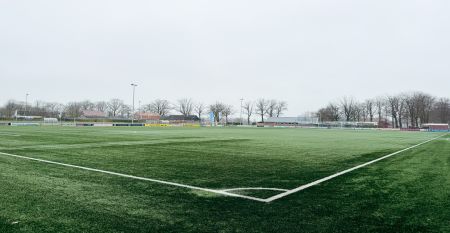 Eine historische Kanone steht auf einem Steinweg im Landschaftsgarten von Marveld Recreatie, vor einem gro&szlig;en Backsteingeb&auml;ude mit modernen Glasfenstern und einem Turm, umgeben von Bungalows unter einem teilweise bew&ouml;lkten Himmel.