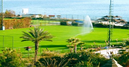 Luftaufnahme des Hotels Limak Arcadia mit Pools, Gärten, Sportplätzen und Strandkabinen - ideal für das Fußballtrainingslager Türkei - inmitten von Palmen und Bergen unter einem klaren Himmel.