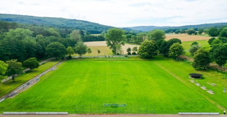 Ein wei&szlig;es, dreist&ouml;ckiges Geb&auml;ude mit braunen Dachziegeln, vielen Fenstern, einer verglasten Terrasse und einem &uuml;ppig belaubten Baum davor. Auf der linken Seite steht auf einem Schild &bdquo;Landhotel Imhof&ldquo;. Dieses familiengef&uuml;hrte Hotel eignet sich ideal f&uuml;r 