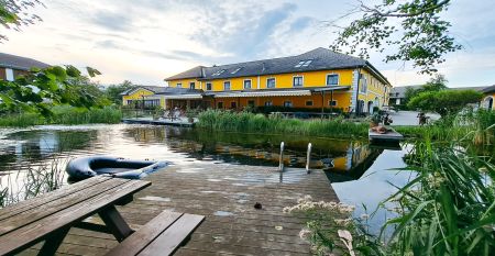 Ein leuchtend gelbes Geb&auml;ude in Neuhofen an der Ybbs mit Steg, Leiter und schwarzem Schlauchboot liegt an einem ruhigen Teich im Gr&uuml;nen - im familiengef&uuml;hrten Hotel Perbersdorfer Heuriger genie&szlig;en die Menschen diese Szene.
