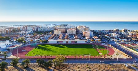 Außenansicht des Hotels Gaudea in Santa Pola mit großen Glasfenstern, die den Blick auf eine moderne, warm beleuchtete Lobby mit einem Sitzbereich freigeben. Der Name des Hotels und die vier Sterne sind über dem Eingang gut sichtbar angebracht.