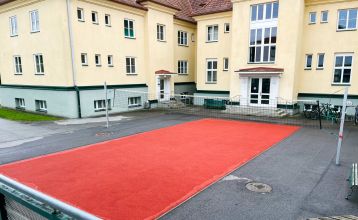 Das Gästehaus Burgenland Güssing ist ein großes, cremefarbenes Gebäude mit rotem Dach und vielen Fenstern unter einem strahlend blauen Himmel, vor dem mehrere Autos geparkt sind und in dessen Nähe Bäume stehen.