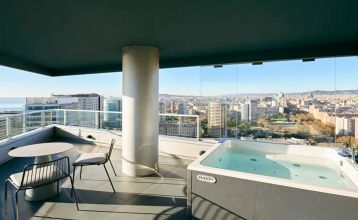 Das Hotel Barcelona Princess ragt inmitten der Hochhäuser Barcelonas nahe der Küste empor, unter einem teilweise bewölkten Himmel, unter dem sich Straßen, Autos und Bäume erstrecken – ein idealer Ort für die sportliche Vorbereitung.
