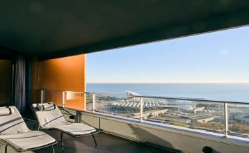Das Hotel Barcelona Princess ragt inmitten der Hochhäuser Barcelonas nahe der Küste empor, unter einem teilweise bewölkten Himmel, unter dem sich Straßen, Autos und Bäume erstrecken – ein idealer Ort für die sportliche Vorbereitung.