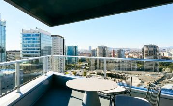 Das Hotel Barcelona Princess ragt inmitten der Hochhäuser Barcelonas nahe der Küste empor, unter einem teilweise bewölkten Himmel, unter dem sich Straßen, Autos und Bäume erstrecken – ein idealer Ort für die sportliche Vorbereitung.