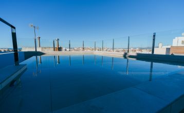 Außenansicht des Hotels Gaudea in Santa Pola mit großen Glasfenstern, die den Blick auf eine moderne, warm beleuchtete Lobby mit einem Sitzbereich freigeben. Der Name des Hotels und die vier Sterne sind über dem Eingang gut sichtbar angebracht.