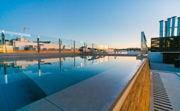 Außenansicht des Hotels Gaudea in Santa Pola mit großen Glasfenstern, die den Blick auf eine moderne, warm beleuchtete Lobby mit einem Sitzbereich freigeben. Der Name des Hotels und die vier Sterne sind über dem Eingang gut sichtbar angebracht.