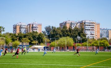 Ein Innenhof mit Palmen und Sitzgelegenheiten im Freien ist von modernen weißen Gebäuden umgeben, mit einem Hotel Zenit Sevilla-Schild - ideal für Fußball-Sportgruppen - und einem großen Gebäude mit der Aufschrift GOOD NIGHT auf dem Dach.