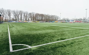 Eine historische Kanone steht auf einem Steinweg im Landschaftsgarten von Marveld Recreatie, vor einem großen Backsteingebäude mit modernen Glasfenstern und einem Turm, umgeben von Bungalows unter einem teilweise bewölkten Himmel.