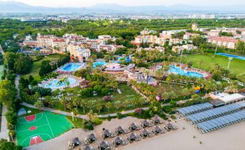 Luftaufnahme des Hotels Limak Arcadia mit Pools, Gärten, Sportplätzen und Strandkabinen - ideal für das Fußballtrainingslager Türkei - inmitten von Palmen und Bergen unter einem klaren Himmel.