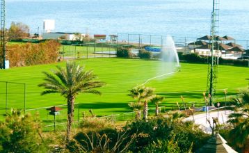 Luftaufnahme des Hotels Limak Arcadia mit Pools, Gärten, Sportplätzen und Strandkabinen - ideal für das Fußballtrainingslager Türkei - inmitten von Palmen und Bergen unter einem klaren Himmel.