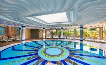 Luftaufnahme des Hotels Limak Arcadia mit Pools, Gärten, Sportplätzen und Strandkabinen - ideal für das Fußballtrainingslager Türkei - inmitten von Palmen und Bergen unter einem klaren Himmel.