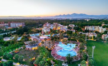 Luftaufnahme des Hotels Limak Arcadia mit Pools, Gärten, Sportplätzen und Strandkabinen - ideal für das Fußballtrainingslager Türkei - inmitten von Palmen und Bergen unter einem klaren Himmel.
