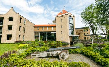 Eine historische Kanone steht auf einem Steinweg im Landschaftsgarten von Marveld Recreatie, vor einem großen Backsteingebäude mit modernen Glasfenstern und einem Turm, umgeben von Bungalows unter einem teilweise bewölkten Himmel.