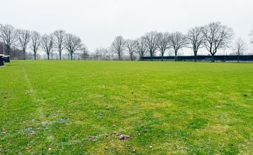 Eine historische Kanone steht auf einem Steinweg im Landschaftsgarten von Marveld Recreatie, vor einem großen Backsteingebäude mit modernen Glasfenstern und einem Turm, umgeben von Bungalows unter einem teilweise bewölkten Himmel.
