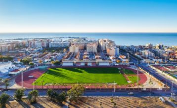 Außenansicht des Hotels Gaudea in Santa Pola mit großen Glasfenstern, die den Blick auf eine moderne, warm beleuchtete Lobby mit einem Sitzbereich freigeben. Der Name des Hotels und die vier Sterne sind über dem Eingang gut sichtbar angebracht.