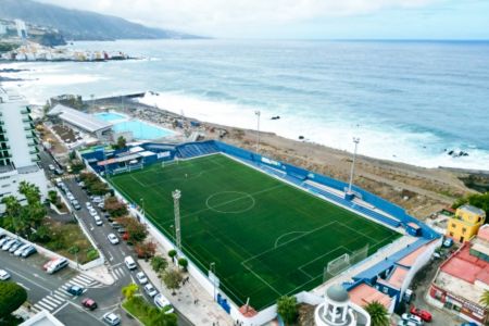 Luftaufnahme der Kunstrasenplätze ElPenon, eines Fußballplatzes direkt am Meer, mit nahegelegenen Schwimmbädern und Gebäuden. Das Stadion liegt in der Nähe einer felsigen Küste, an der die Wellen gegen das Ufer branden; links ist eine Stadt zu sehen.