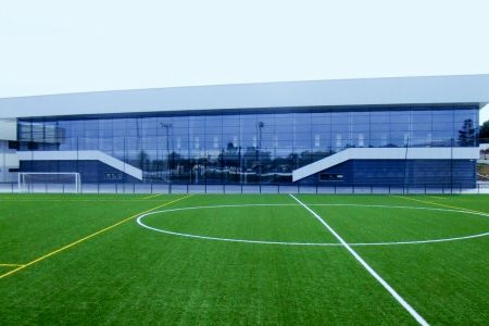 Im Vordergrund befindet sich der Kunstrasenplatz Altea, ein Fußballplatz mit weiß-gelben Markierungen, während ihm gegenüber ein modernes Gebäude mit großen Glasfenstern steht.