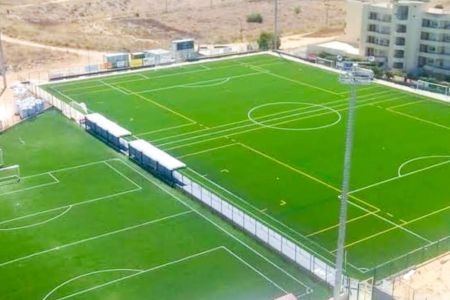 Luftaufnahme des „Campo David Neto N2“ in Portimão, auf der zwei nebeneinander liegende Fußballplätze mit Kunstrasen und weißen sowie gelben Spielfeldmarkierungen zu sehen sind, die von einem Zaun umgeben sind; im Hintergrund sind Gebäude und trockenes La
