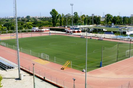 Luftaufnahme des Hauptplatzes Oliva mit mehreren Gebäuden, einem Fußballplatz, einer Laufbahn, Tennisplätzen und einem Parkplatz inmitten von Grünanlagen mit einer Stadt im Hintergrund.