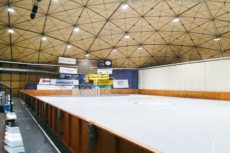 Die Sporthalle Hollabrunn ist eine leere Sporthalle mit einer kuppelförmigen Decke, Holzbande rund um das Spielfeld, Werbebannern an der gegenüberliegenden Wand und leeren Tribünen auf der linken Seite.
