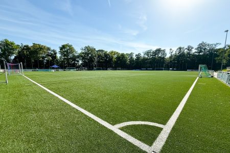 Ein sonniger Tag auf dem Kunstrasenplatz Schöndorf: ein leerer Fußballplatz im Freien mit grünem Kunstrasen, strahlend weißen Begrenzungslinien, zwei Toren, Bäumen im Hintergrund und einem strahlend blauen Himmel über den Köpfen.