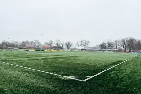 Weitblick auf den leeren, grünen Fußballplatz des SV Grol Kunstrasen 2 an einem bewölkten Tag, mit weißen Feldlinien und Bäumen, Gebäuden und Zäunen im Hintergrund.