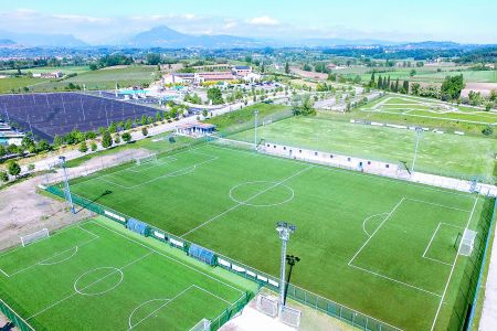 Luftaufnahme des Kunstrasenplatzes Lazise, einer weitläufigen Sportanlage mit mehreren grünen Fußballplätzen, Parkplätzen und Gebäuden, eingebettet zwischen Bäumen und ländlicher Idylle unter strahlend blauem Himmel.