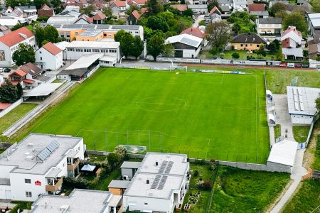 Luftaufnahme des Naturrasenplatzes Horitschon, eines grünen Fußballplatzes mit Tribünen auf einer Seite und Torpfosten an beiden Enden, umgeben von Wohngebäuden und Bäumen in einem Vorstadtbezirk.