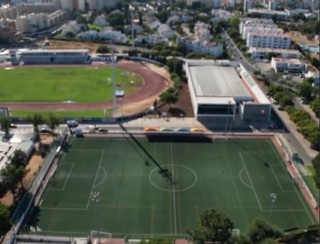 Luftaufnahme des Estadio Municipal Quarteria N1, im Vordergrund ein Fußballfeld mit grünem Kunstrasen und im Hintergrund eine Laufbahn mit Rasenfläche, alles umgeben von Gebäuden und Bäumen.