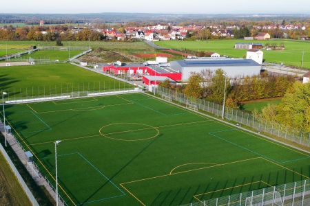 Luftaufnahme von zwei eingezäunten Kunstrasen-Fußballplätzen im Roßtal, umgeben von Gebäuden, Bäumen, offenen Grasflächen und einer kleinen Stadt im Hintergrund.