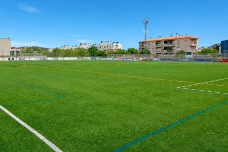 Weite Sicht auf das leere Camp Municipal de Futbol Vilartagues mit grünem Kunstrasen, weißen und gelben Linien und Gebäuden im Hintergrund unter einem klaren blauen Himmel.