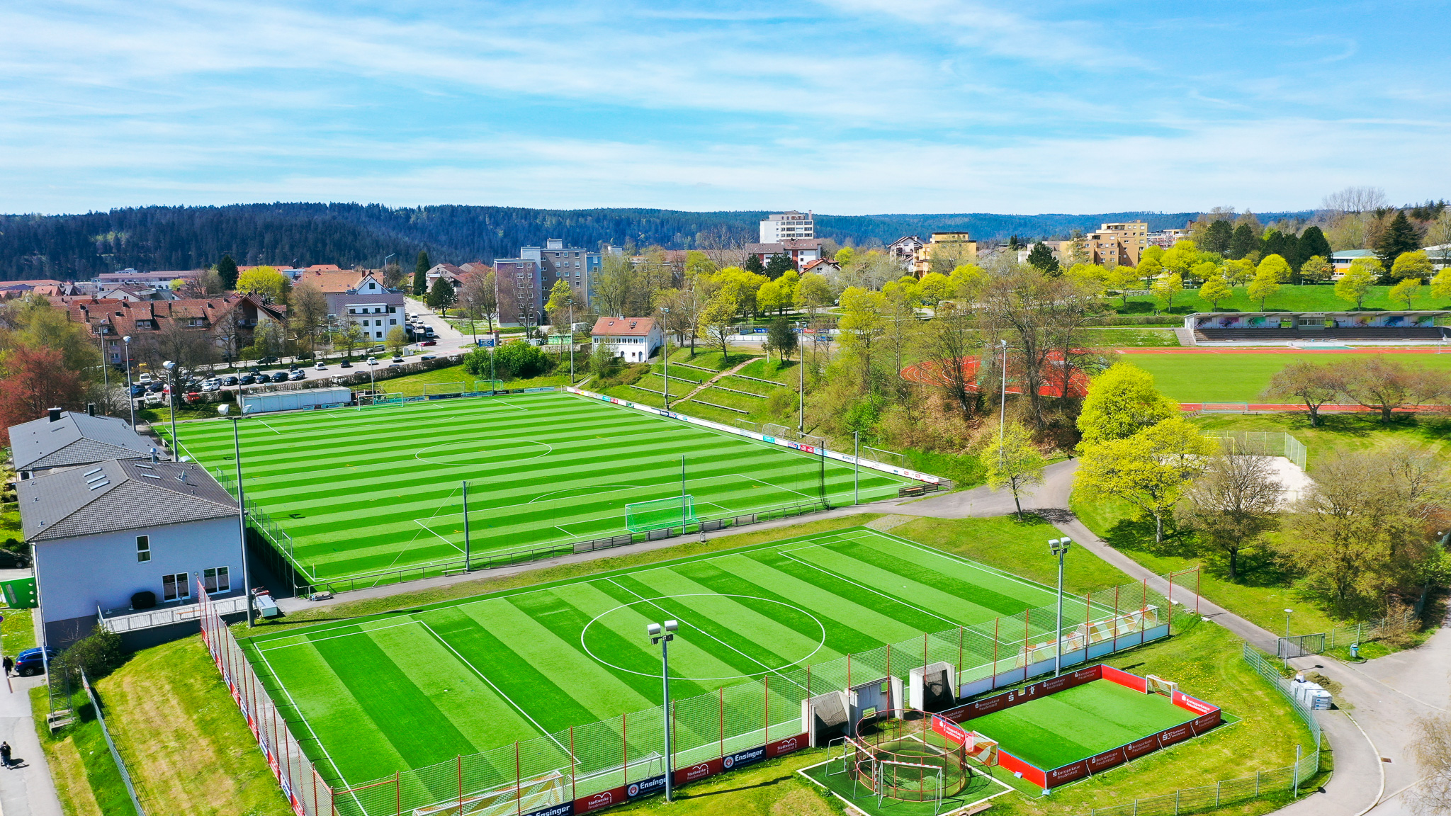 Luftaufnahme der Fußballtrainingsanlage in Baden-Württemberg mit zwei Kunstrasenplätzen, Kleinfeld, Flutlicht und Tribüne, eingebettet in eine grüne Umgebung.