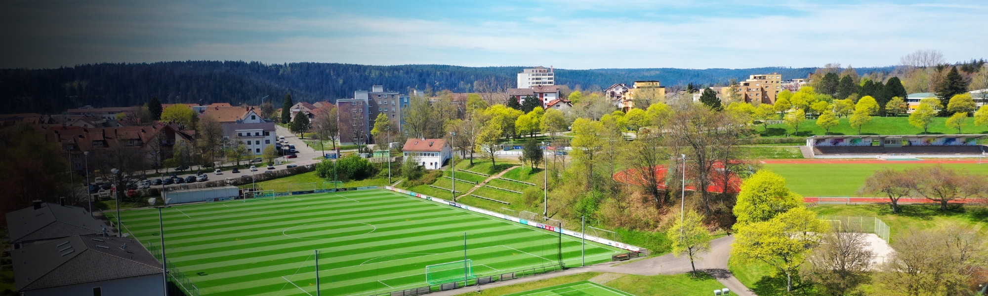 Luftaufnahme einer modernen Sportanlage in Baden-Württemberg mit mehreren Kunstrasenplätzen, einer Leichtathletikanlage und umgeben von Bäumen und Wohngebäuden.