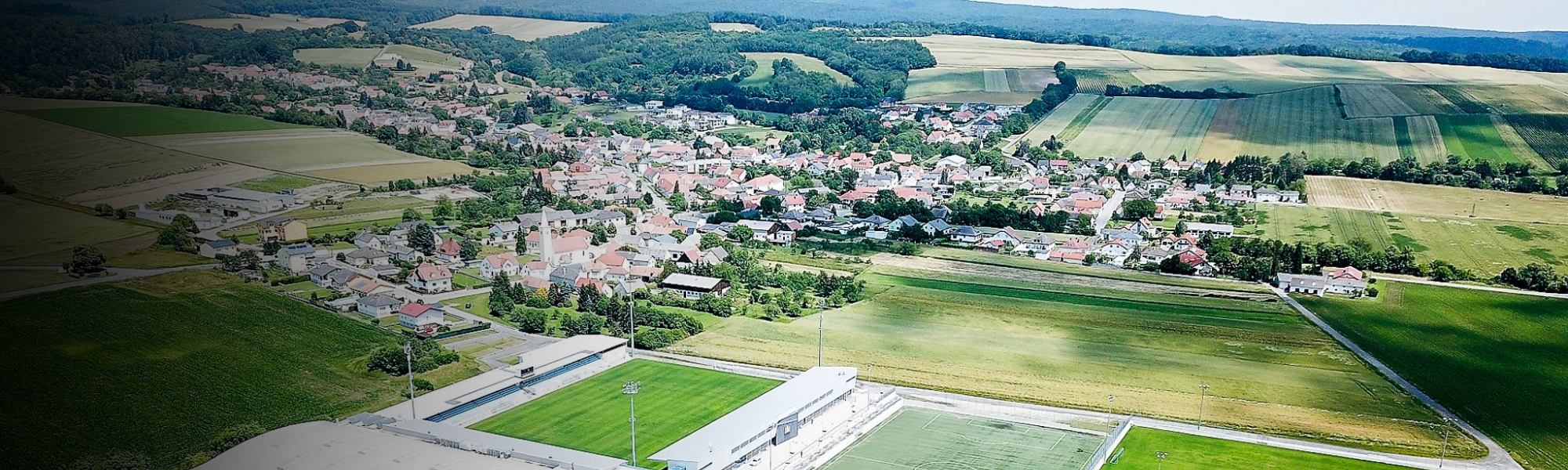 Luftaufnahme einer Sportanlage im Burgenland, umgeben von grünen Feldern und einem kleinen Dorf mit roten Dächern.