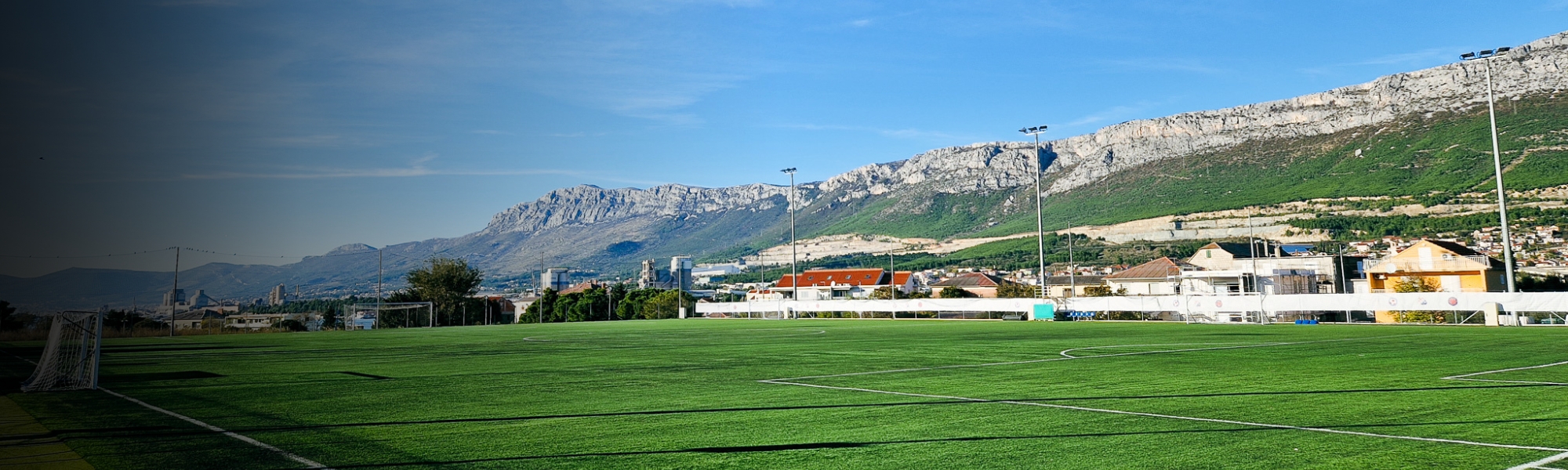 Fußballplatz in Dalmatien mit Flutlichtmasten und beeindruckendem Bergpanorama im Hintergrund, unter klarem, blauem Himmel.