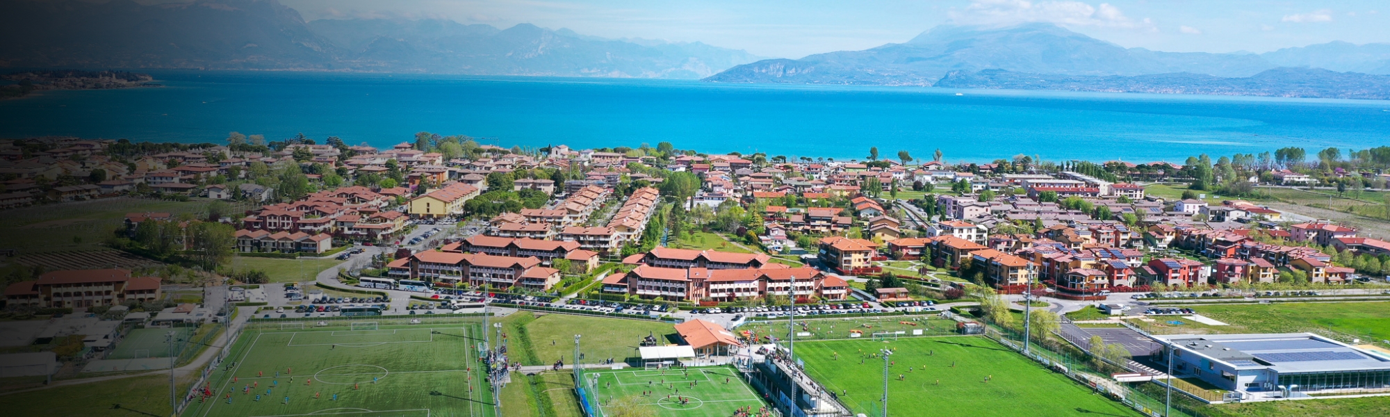 Panoramaaufnahme einer Sportanlage mit mehreren Fußballplätzen am Gardasee, eingerahmt von Weinbergen, Wohnhäusern und dem See im Hintergrund.