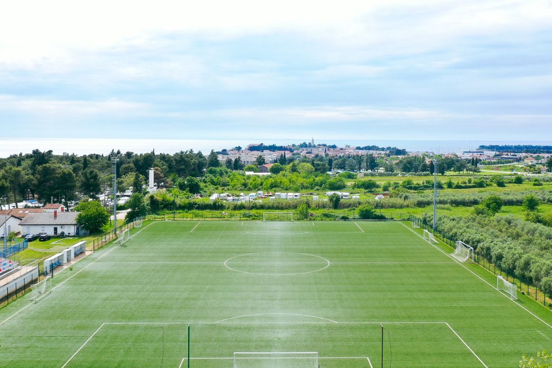  Fußballplatz in Istrien mit Kunstrasen, umgeben von grüner Natur und Olivenbäumen, im Hintergrund das Meer und die Stadt Poreč.