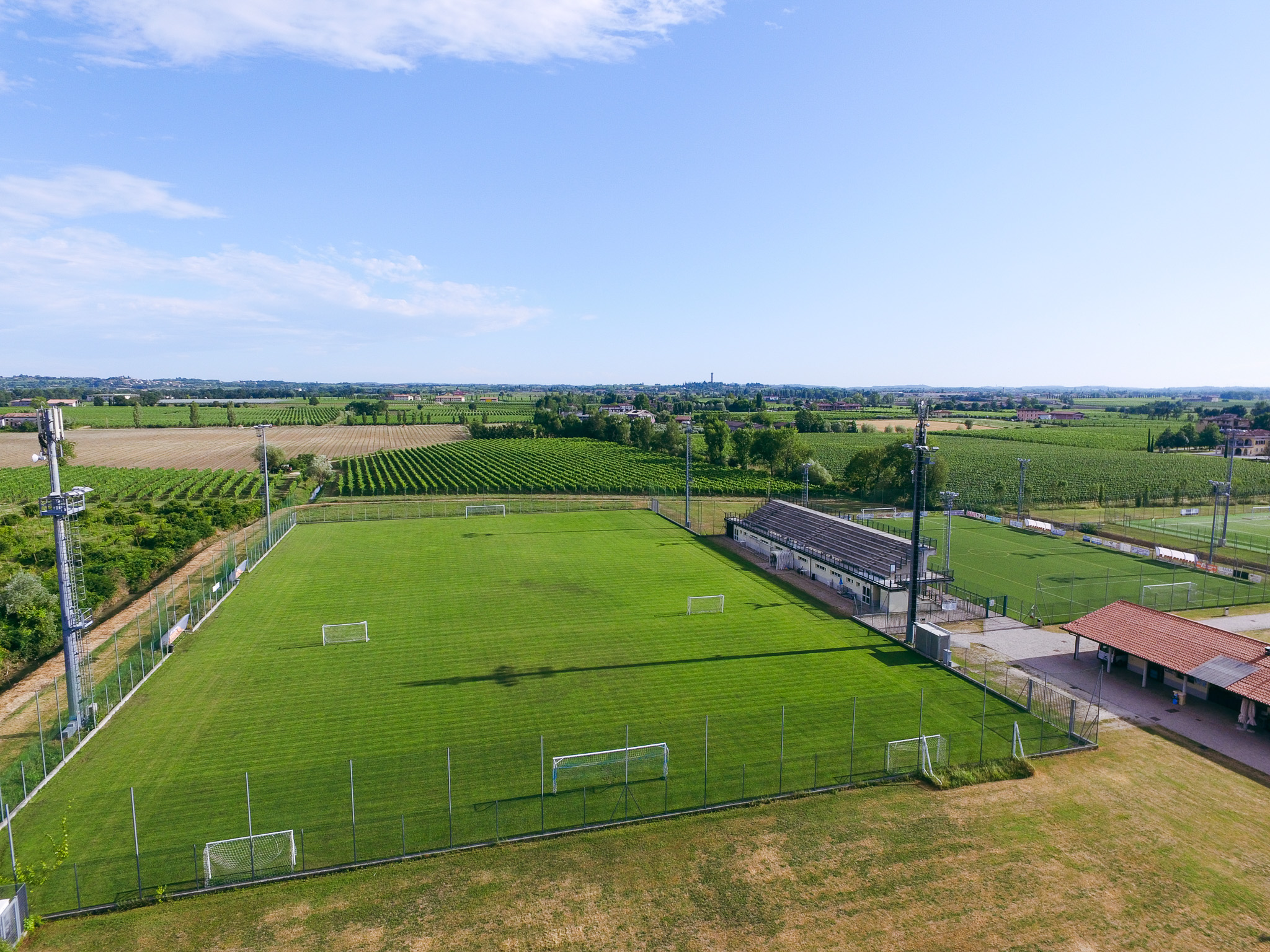 Luftaufnahme einer Sportanlage mit mehreren Fußballplätzen, umgeben von einer italienischen Kleinstadt am Ufer eines Sees, mit Bergen im Hintergrund.