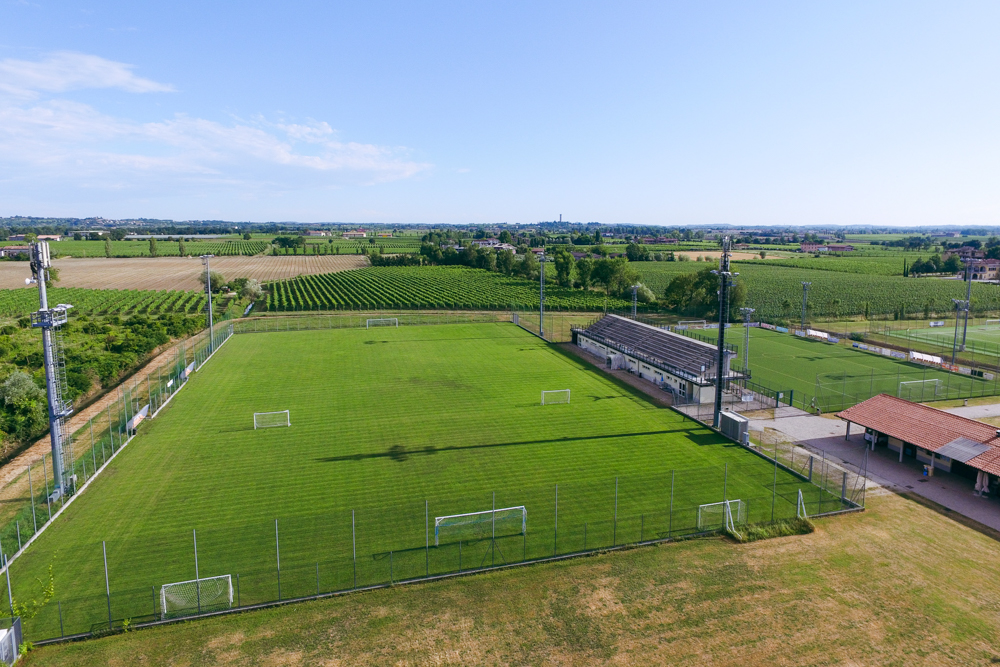 Fußballtrainingsplatz auf Sardinien mit grünem Kunstrasen, Tor, Bänken und Flutlichtmasten, umgeben von Netzen und Berglandschaft.