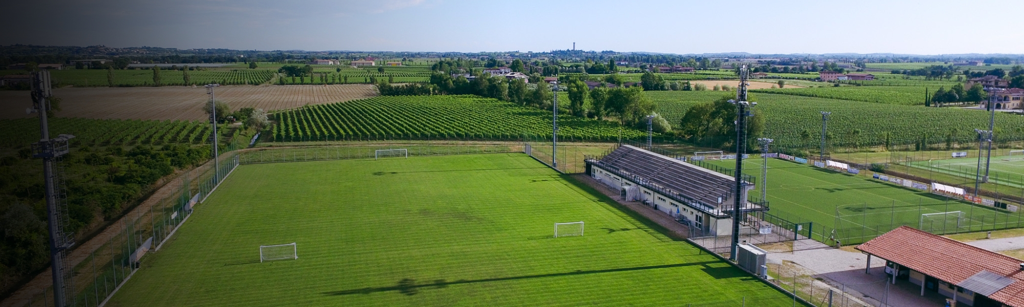 Panorama-Luftaufnahme einer Sportanlage mit mehreren Fußballplätzen, umgeben von einer italienischen Kleinstadt am Ufer eines Sees.