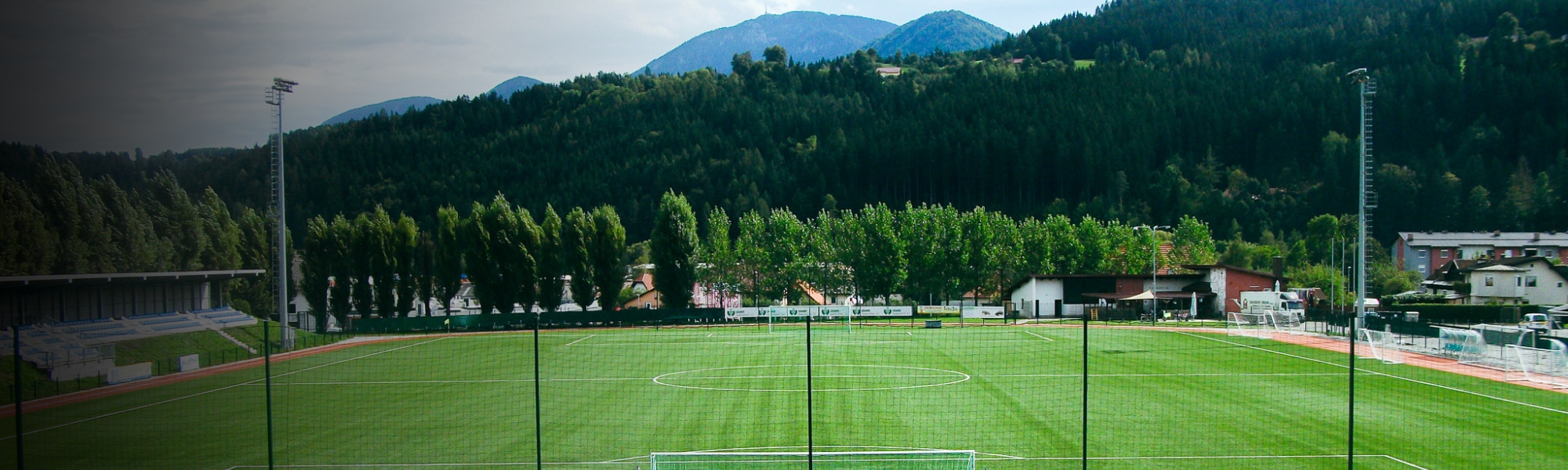 Fußballplatz in Kärnten, umgeben von grünen Wäldern und Berglandschaft, mit moderner Tribüne und Flutlichtanlage.