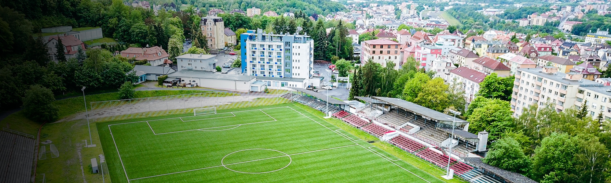 Fußballstadion mit Tribünen, umgeben von einer hügeligen Stadtlandschaft mit vielen Bäumen.