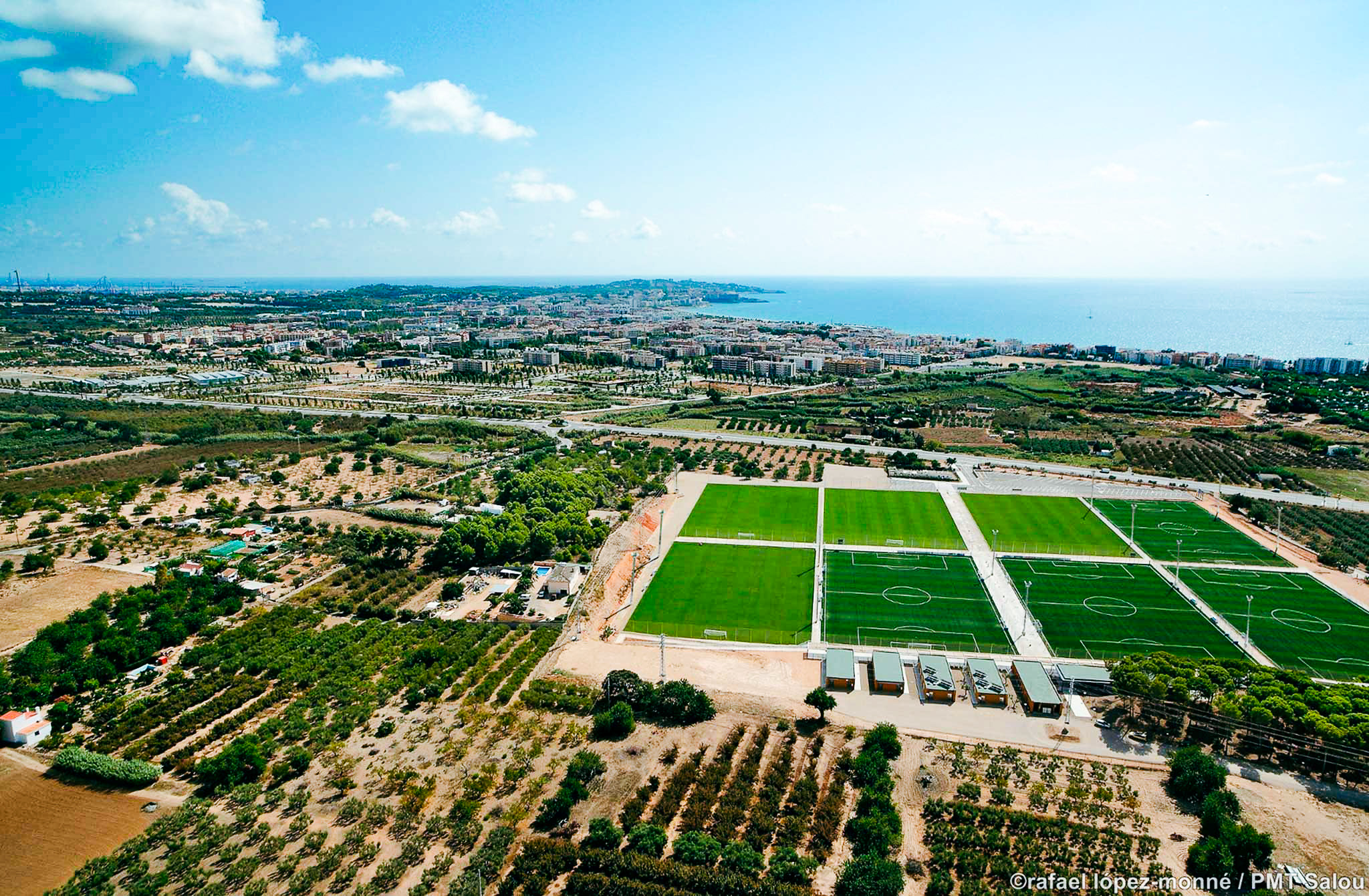 Eine moderne Sportanlage mit mehreren Fußballfeldern, Küstenaussicht und urbaner Umgebung.