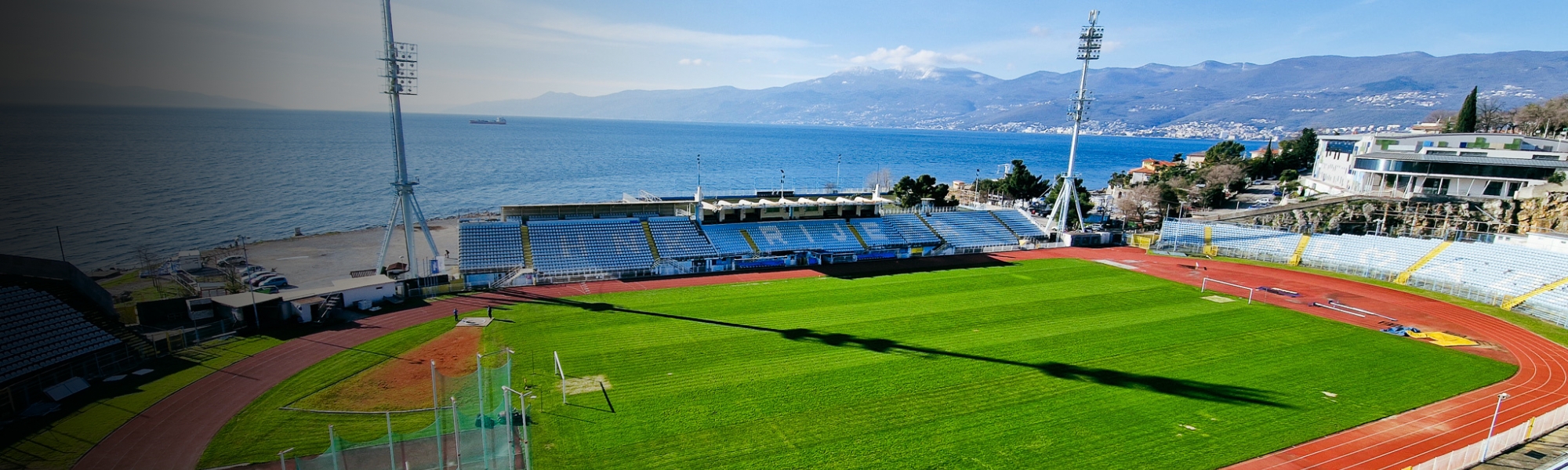Panoramaansicht eines Fußballstadions an der Kvarner Bucht, mit Blick auf das Spielfeld, die Laufbahn, die blauen Tribünen und das glitzernde Meer, eingerahmt von einer bergigen Küstenlandschaft.