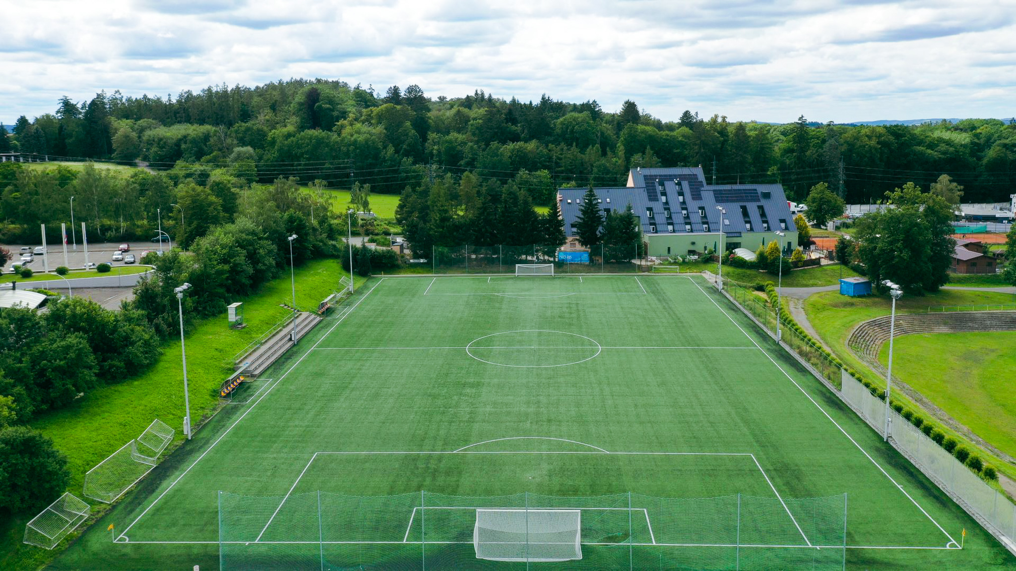 Fußballplatz mit Kunstrasen in einer idyllischen Umgebung, angrenzend an ein modernes Gebäude.